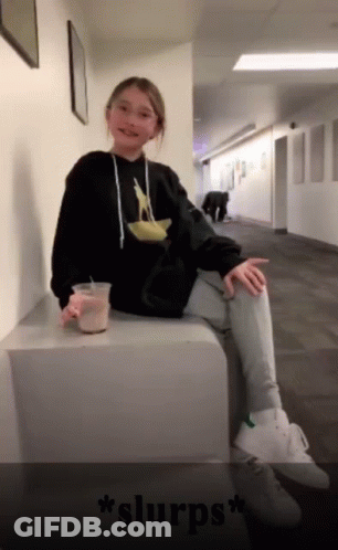 Girl In Black Hoodie Sitting On Cube Holding Coffee GIF