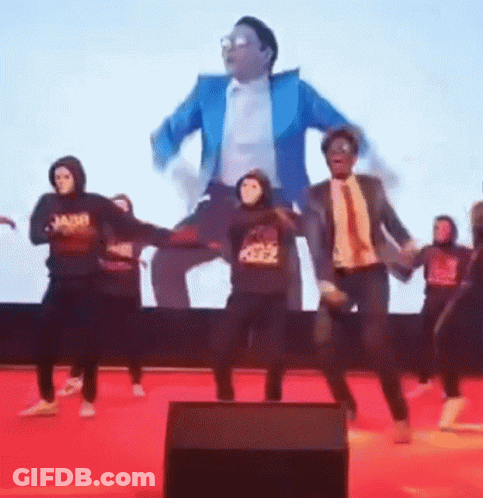 Group Dancing In Masks In Front Of Tuxedo Man On Screen GIF