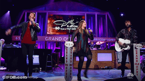 Group Singing And Playing Guitars Behind Dolly Parton Screen GIF