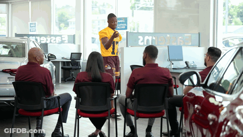 Group Sitting In Car Showroom With Man In Yellow Shirt Number 9 GIF