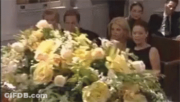 Group Sitting In Front Of Coffin Covered In Flowers GIF