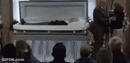 Group Sitting In Front Of Coffin With Man Standing GIF