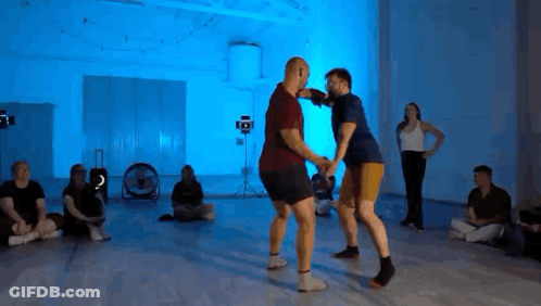 Group Sitting On Floor With Blue Lights While Two Men Dance GIF