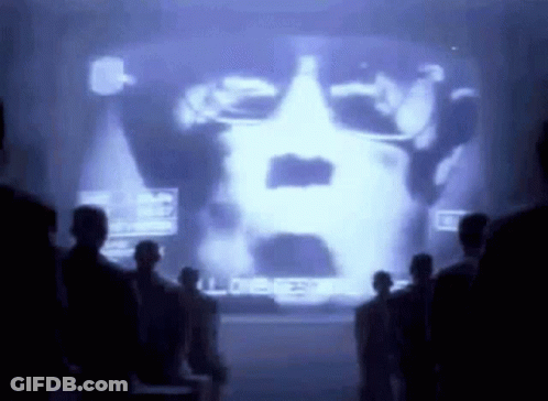 Group Standing Before Large Screen Showing Man's Face GIF