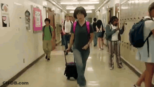 Group Walking Down School Hallway With Woman Pulling Suitcase GIF