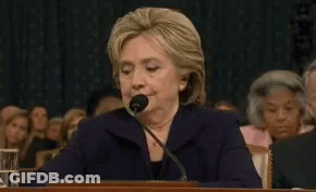 Hillary Clinton Is Speaking Into A Microphone While Sitting At A Desk In Front Of A Crowd . GIF