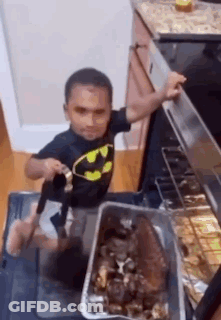 Little Boy In Batman Shirt Standing By Oven With Pan Of Food GIF