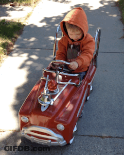 Little Boy In Orange Hoodie Sitting In Red Toy Car GIF