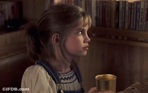 Little Girl Holding Cup In Front Of Bookshelf GIF