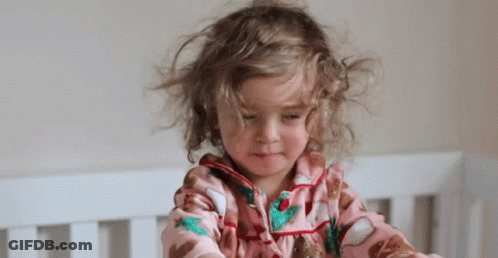 Little Girl With Curly Hair Sitting In Crib Looking At Camera GIF