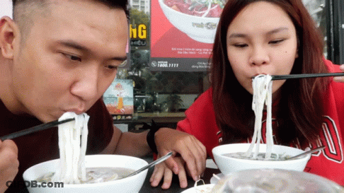 Man And Woman Eating Noodles With Chopsticks In Front Of Ung Sign GIF