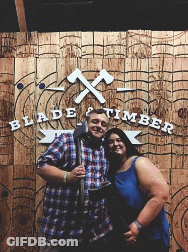 Man And Woman Posing In Front Of Blade And Timber Sign GIF