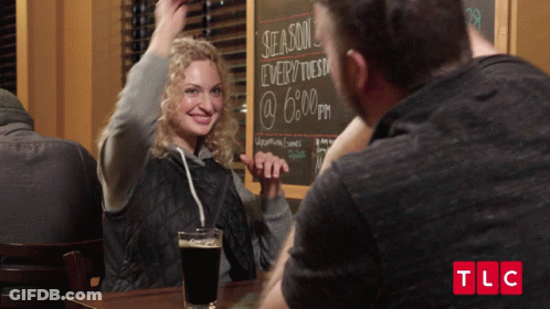Man And Woman Sitting At Table With Chalkboard Saying Season Every Tuesday @ 6 PM GIF