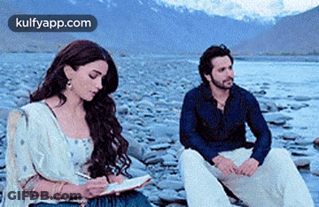 Man And Woman Sitting On Rocky Beach Woman Writing In Notebook GIF