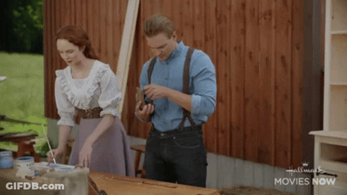 Man And Woman Standing Beside Hallmark Movies Now Table GIF