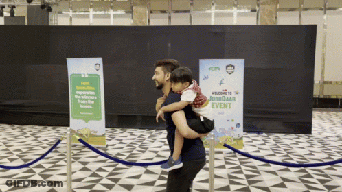 Man Carrying Child In Front Of Welcome To Jorbadar Event Sign GIF
