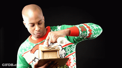 Man Decorating Gingerbread House In Green And Red Sweater GIF