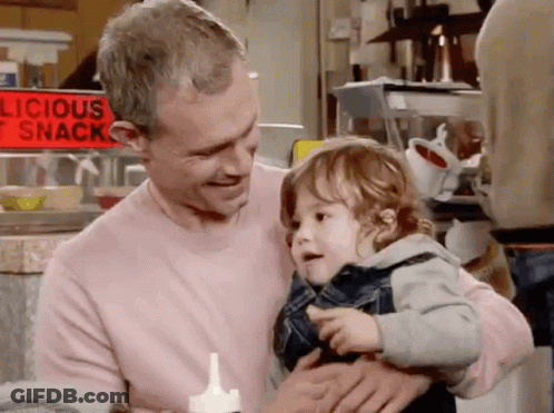 Man Holding Baby In Front Of Delicious Snack Sign GIF