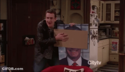 Man Holding Cardboard Box With Man Picture In Front Of Citytv Refrigerator GIF