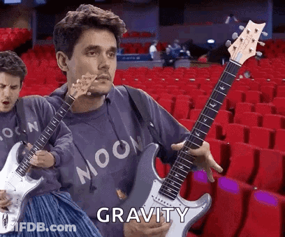 Man Holding Guitar In Front Of Red Seats Says Gravity GIF