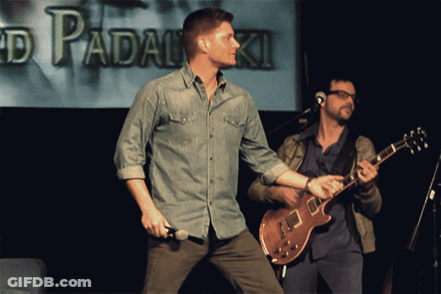 Man Holding Microphone Next To Guitarist In Front Of Padalski Screen GIF