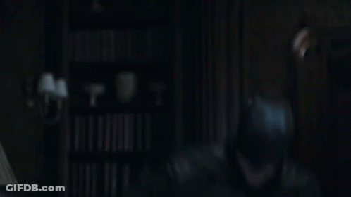 Man In Batman Costume In Dark Room In Front Of Bookshelf GIF