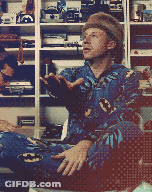 Man In Batman Pajama Sitting In Front Of Camera Shelves GIF