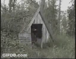 Man In Black And White Photo Holding Gun In Front Of Wooden Building GIF