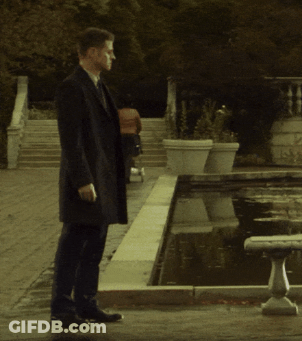 Man In Black Coat Walking By Fountain GIF