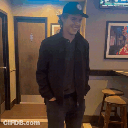 Man In Black Hat Smiling In Front Of Bathroom Door GIF