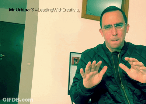 Man In Black Jacket Sitting In Front Of Mr Urbina #LeadingWithCreativity Sign GIF