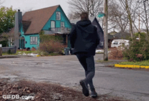 Man In Black Jacket Walking Past Blue House GIF