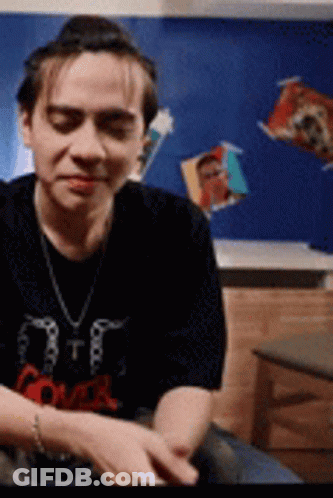 Man In Black Shirt And Necklace Sitting By Blue Wall GIF