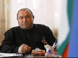 Man In Black Shirt At Desk With Pen Holder In Front Of Flag GIF