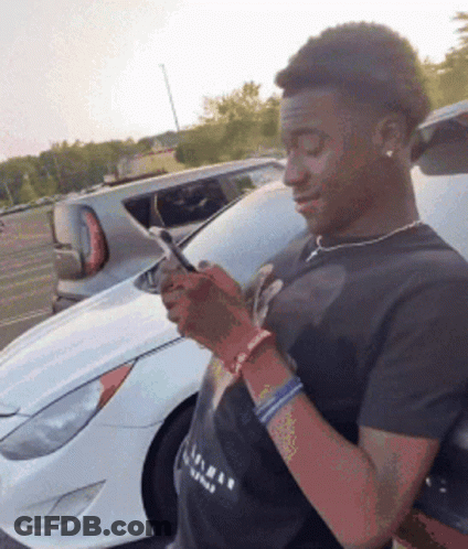 Man In Black Shirt Looking At Phone Next To White Car GIF