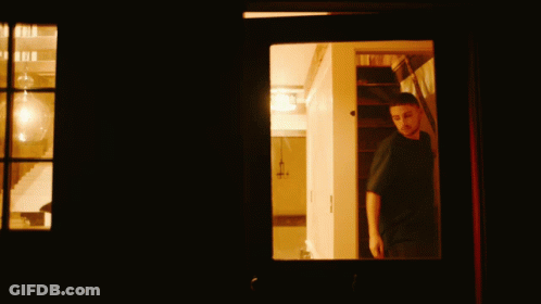 Man In Black Shirt Looking Out Window In Dark Room GIF
