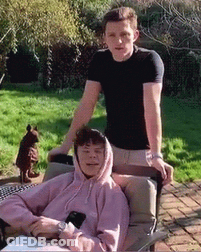 Man In Black Shirt Standing Next To Man In Pink Hoodie Sitting On Chair GIF