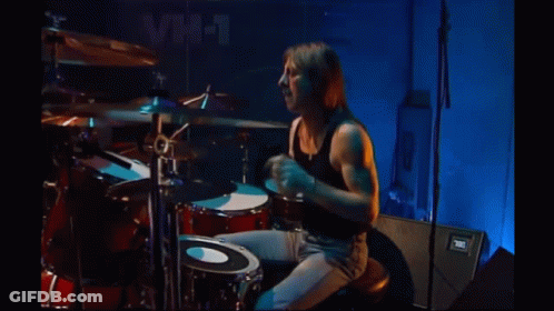 Man In Black Tank Top Playing Drums In Front Of VH-1 Sign GIF