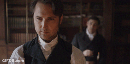 Man In Black Vest And White Shirt In Front Of Bookshelf GIF