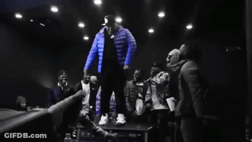 Man In Blue Jacket Standing On Table In Front Of Crowd GIF