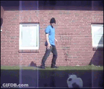 Man In Blue Shirt And Black Hat Walking By Brick Building GIF