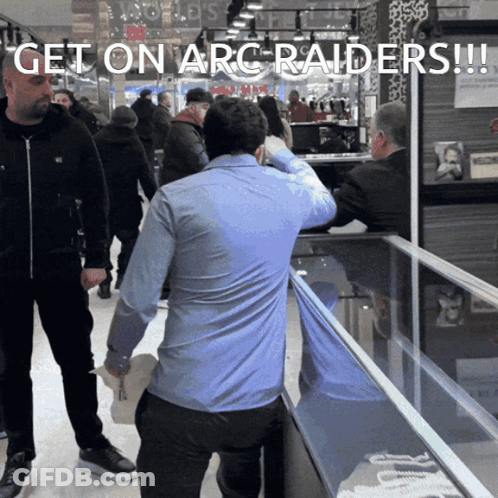 Man In Blue Shirt In Front Of Get On Arc Raiders Display Case GIF