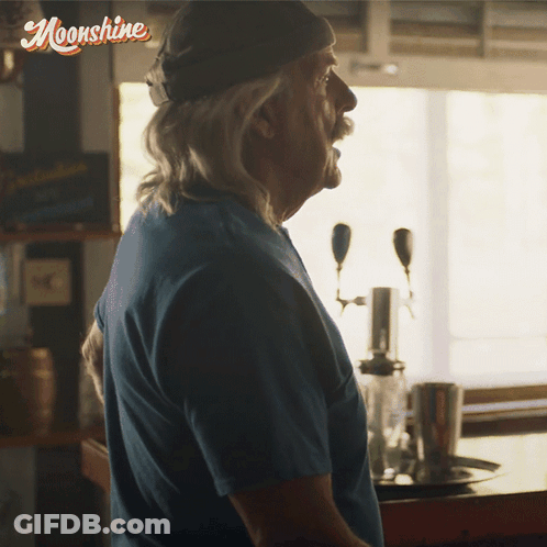 Man In Blue Shirt In Front Of Moonshine Sign GIF