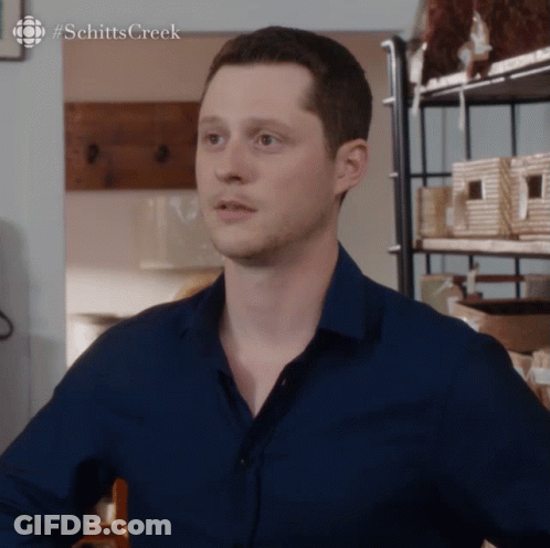 Man In Blue Shirt In Front Of Schitts Creek Shelf GIF