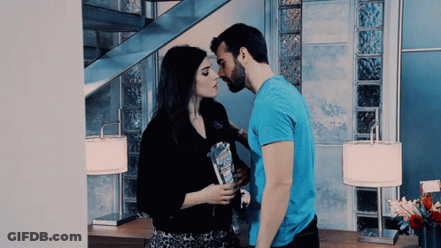 Man In Blue Shirt Kissing Woman On Cheek In Front Of Staircase GIF