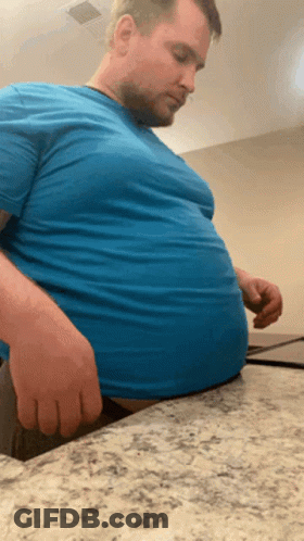 Man In Blue Shirt Sitting On Counter With Hands On Stomach GIF