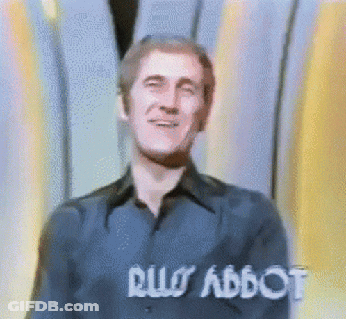 Man In Blue Shirt Smiling In Front Of Rus Abbot Sign GIF