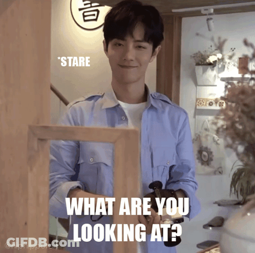 Man In Blue Shirt Standing Before What Are You Looking At Sign GIF