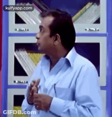 Man In Blue Shirt Standing In Front Of Bookshelf With Hands In Pockets GIF