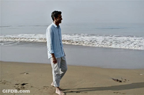 Man In Blue Shirt Walking Barefoot On Beach GIF